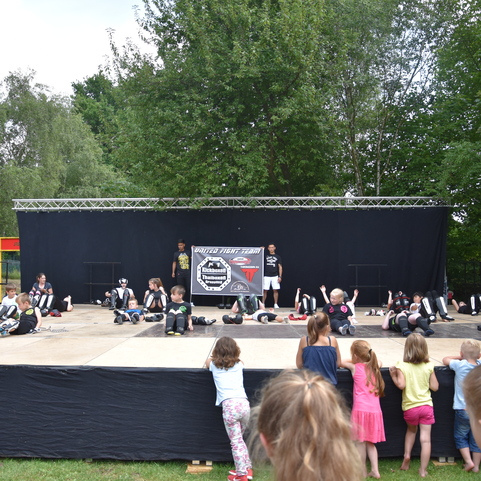Gruppenvorf&uuml;hrung von Kindern auf einer B&uuml;hne, Zuschauer stehen davor, B&auml;ume im Hintergrund.