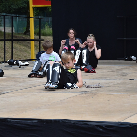 Gruppe von Kindern und Jugendlichen in Schutzausr&uuml;stung sitzt auf einer Holzplattform w&auml;hrend einer Sporteinheit.