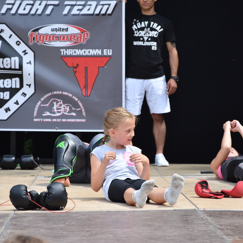 M&auml;dchen mit Kopfschutz sitzt auf dem Boden eines Sportgel&auml;ndes, w&auml;hrend andere Kinder &Uuml;bungen machen. Hintergrund mit Banner.