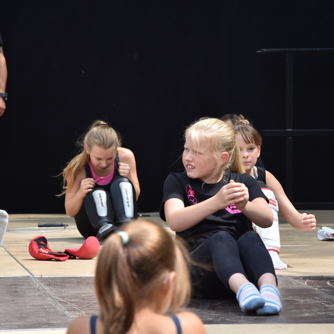 Kinder in Trainingstops f&uuml;hren &Uuml;bungen auf einer Sportfl&auml;che durch, w&auml;hrend ein Trainer im Hintergrund zuschaut.