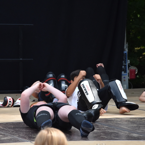 Kinder in Schutzausr&uuml;stung &uuml;ben am Boden liegend Technikbewegungen auf einer Kampfsportmatte.