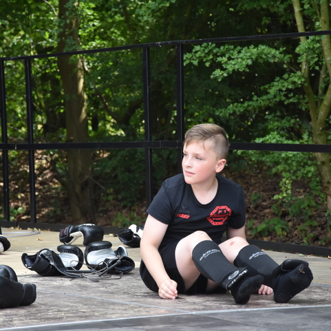 Zwei Kinder in Kampfsportausr&uuml;stung sitzen auf einem Holzuntergrund, umgeben von B&auml;umen und gr&uuml;nem Laub.