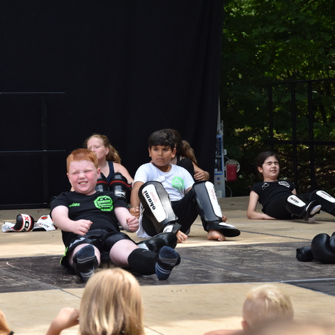 Kinder in Trainingskleidung sitzen auf einer Turnfl&auml;che, w&auml;hrend sie mit Schutzausr&uuml;stung f&uuml;r Kampfsport &uuml;ben.