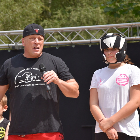 Ein Mann spricht zu einer Gruppe, w&auml;hrend ein M&auml;dchen mit Schutzhelm neben ihm steht. Im Hintergrund stehen weitere Kinder.