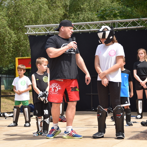 Trainer erkl&auml;rt und demonstriert eine Technik f&uuml;r Kinder in Schutzausr&uuml;stung w&auml;hrend eines Kampfsport-Events im Freien.