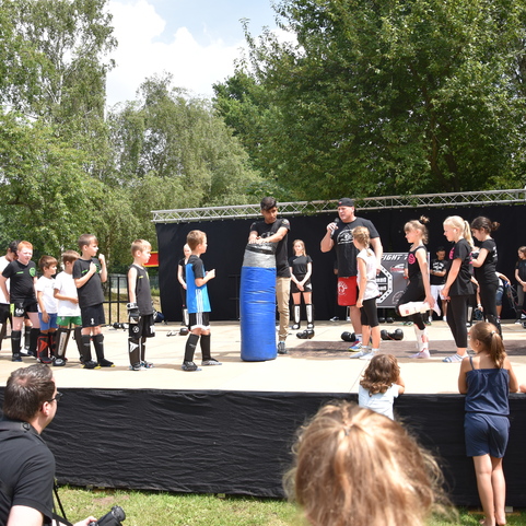 Kinder und Trainer stehen um einen blauen Boxsack auf einer B&uuml;hne, umringt von Zuschauern bei einer Sportveranstaltung.