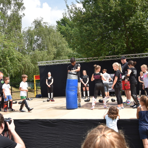 Gruppentraining mit Kindern an einer B&uuml;hne, auf der Techniken am Boxsack demonstriert werden. Zuschauer beobachten aufmerksam.