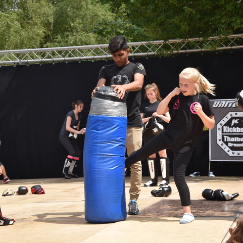 M&auml;dchen tritt gegen einen blauen Boxsack, w&auml;hrend ein Trainer unterst&uuml;tzt; im Hintergrund weitere Kinder in Training.