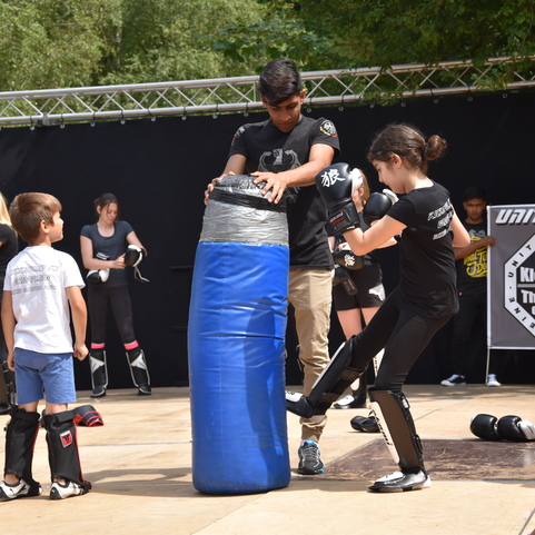 Kinder und Jugendliche trainieren Kickboxen, w&auml;hrend ein Junge einen blauen Trainingssack h&auml;lt und ein M&auml;dchen einen Kick ausf&uuml;hrt.