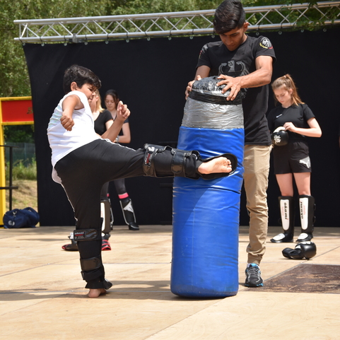 Ein Kind tritt mit einem Bein gegen einen Trainingssack, w&auml;hrend ein Trainer Anweisungen gibt; andere Kinder beobachten.