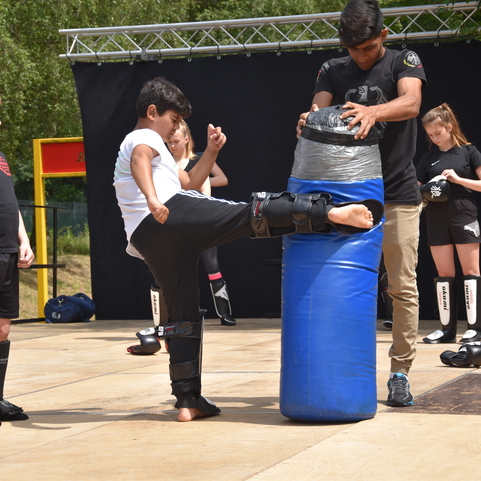 Ein Junge mit Sch&uuml;tzer tritt gegen ein blauer Schlagpolster, w&auml;hrend andere Kinder zuschauen.