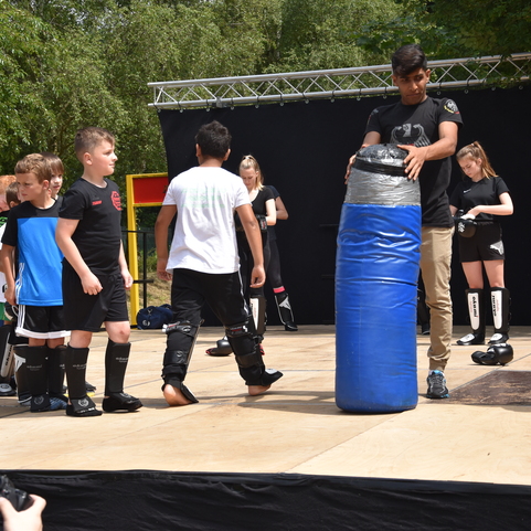 Kinder in Sportkleidung stehen auf einer B&uuml;hne, w&auml;hrend ein Trainer einen blauen Schlagpolster pr&auml;sentiert.