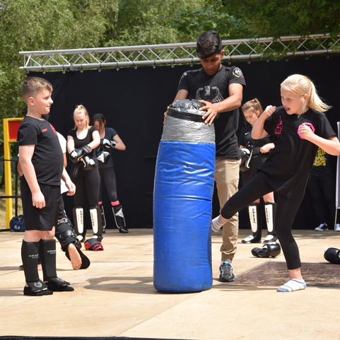 Kinder &uuml;ben Kickboxen, w&auml;hrend ein Junge einen blauen Sandsack h&auml;lt und ein M&auml;dchen mit dem Fu&szlig; darauf tritt.