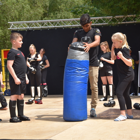 Eine Gruppe von Kindern beobachtet einen Trainer, der einen blauen Boxsack auf einer Trainingsfl&auml;che h&auml;lt.