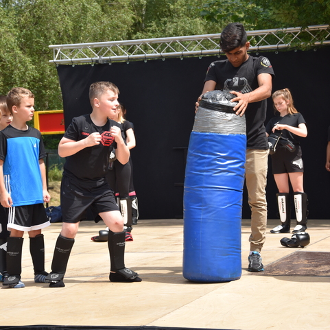 Kinder in Sportkleidung &uuml;ben Kamfsporttechniken an einem blauen Trainingssack auf einer B&uuml;hne im Freien.