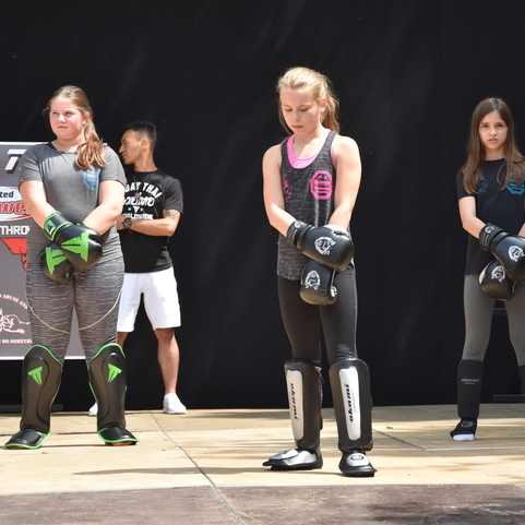 Drei junge K&auml;mpferinnen in Schutzausr&uuml;stung stehen mit F&auml;usten zum Training bereit, im Hintergrund ein Trainer.