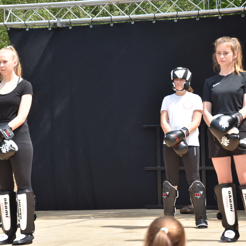 Drei Personen in Sportkleidung stehen auf einer B&uuml;hne, zwei tragen Boxhandschuhe und Schutzausr&uuml;stung.