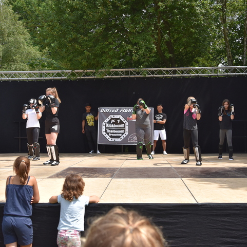 Eine Gruppe von acht Sportlern in Boxhandschuhen &uuml;bt auf einer B&uuml;hne, Zuschauer stehen am Rand.