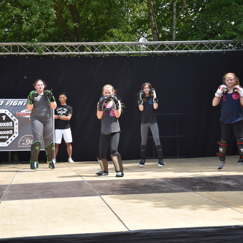 Sechs Jugendliche trainieren Kickboxen auf einer B&uuml;hne, ausgestattet mit Handschuhen und Schutzausr&uuml;stung.