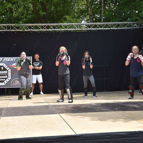 M&auml;dchen in Kampfsportausr&uuml;stung &uuml;ben Kickboxen auf einer B&uuml;hne, umgeben von Zuschauern und einem Banner im Hintergrund.