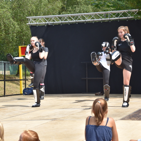 Drei Kampfsportlerinnen f&uuml;hren Kicks auf einer B&uuml;hne vor Publikum w&auml;hrend einer Vorf&uuml;hrung aus.