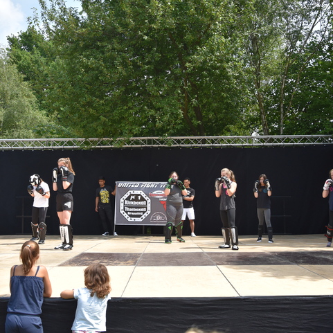 Gruppe von K&auml;mpfern in Schwarz auf einer B&uuml;hne beim Training, beobachtet von Jugendlichen im Hintergrund.