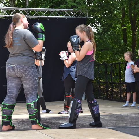 Zwei M&auml;dchen im Sparring mit Schutzausr&uuml;stung und Handschuhen auf einer Trainingsfl&auml;che, umgeben von Zuschauern.