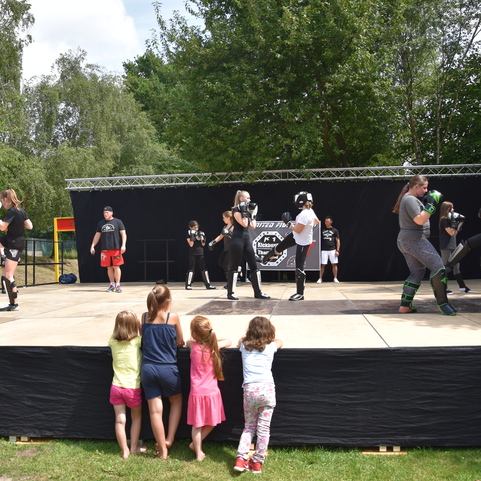 Zwei Gruppen von K&auml;mpfern trainieren gemeinsam in einer Indoor-Arena, w&auml;hrend Kinder am Rand zuschauen.
