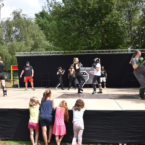 Gruppe von K&auml;mpfern in Sportausr&uuml;stung auf einer B&uuml;hne, w&auml;hrend Kinder ihnen zuschauen. Die Kulisse zeigt B&auml;ume im Hintergrund.