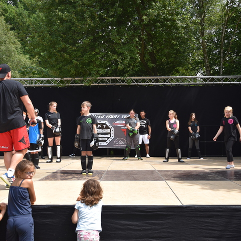 Blick auf eine Trainingssession mit Kindern in Sportkleidung auf einer B&uuml;hne, &uuml;berwacht von einem Trainer in schwarzem Shirt.