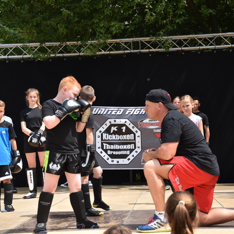 Ein Trainer zeigt einem jungen K&auml;mpfer die richtige Technik beim Kickboxen auf einer B&uuml;hne vor Zuschauern.