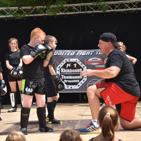 Trainer zeigt Kindern im Kampfanzug eine Technik im Kickboxen w&auml;hrend einer Vorf&uuml;hrung. Zuschauer beobachten aufmerksam.