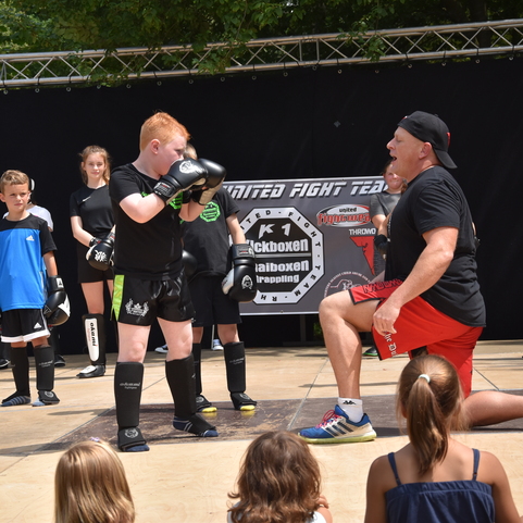 Zwei Kinder im Box-Outfit trainieren auf einer B&uuml;hne, w&auml;hrend ein Trainer Anweisungen gibt. Publikum schaut zu.