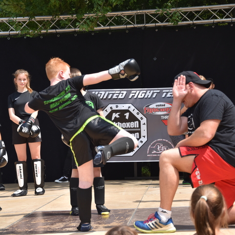 Junge im schwarzen Kampfanzug f&uuml;hrt einen Kick gegen einen Trainer auf einer Kampfsportveranstaltung aus. Zuschauer im Hintergrund.