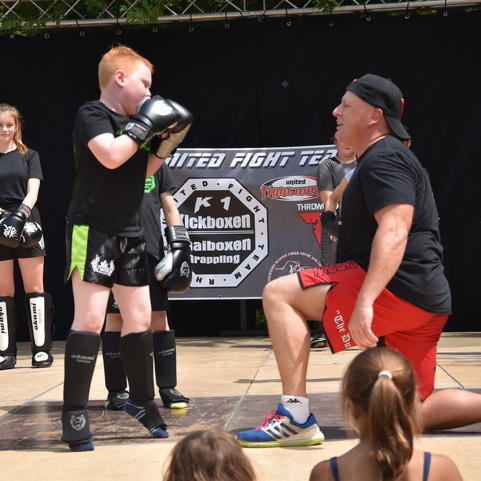 Ein Trainer kniet vor einem Jungen, der Boxhandschuhe tr&auml;gt und einen Schlag ausf&uuml;hrt, umgeben von anderen Kindern.