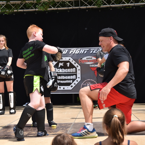 Kind im Training, das einem Trainer mit Handschuhen eine Technik zeigt, w&auml;hrend Zuschauer im Hintergrund beobachten.