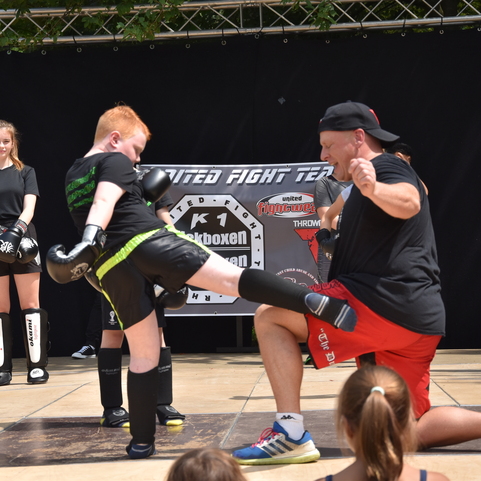 Ein Kind tritt gegen ein gepolstertes Kissen, w&auml;hrend ein Trainer kniet und Anweisungen gibt, umgeben von weiteren Kindern.