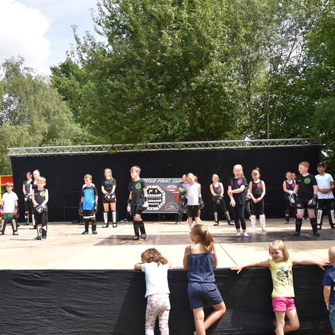 Gruppentraining f&uuml;r Kinder auf einer B&uuml;hne, angeleitet von einem Trainer, im Freien mit B&auml;umen im Hintergrund.