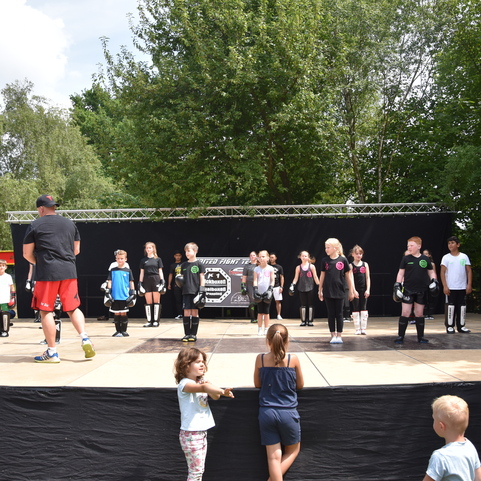 Kinder &uuml;ben in Sportkleidung auf einer B&uuml;hne unter Anleitung eines Trainers, umgeben von B&auml;umen und Sonnenschein.