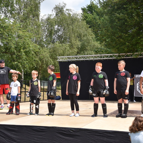 Gruppe von Kindern in Sportkleidung mit Boxhandschuhen stehen auf einer B&uuml;hne w&auml;hrend einer Sportveranstaltung im Freien.