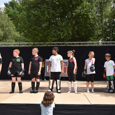 Gruppe von Kindern in Boxausr&uuml;stung steht auf einer B&uuml;hne, bereit f&uuml;r eine Vorf&uuml;hrung, vor einem schwarzen Hintergrund.