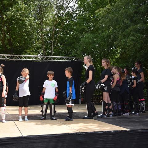 Gruppentraining von Kindern in Sportkleidung mit Boxhandschuhen auf einer B&uuml;hne im Freien.