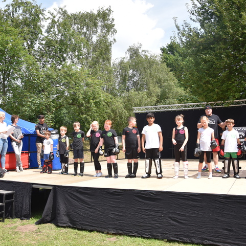 Gruppe von Kindern in Sportbekleidung steht auf einer B&uuml;hne, w&auml;hrend Zuschauer auf einer Wiese zuschauen.