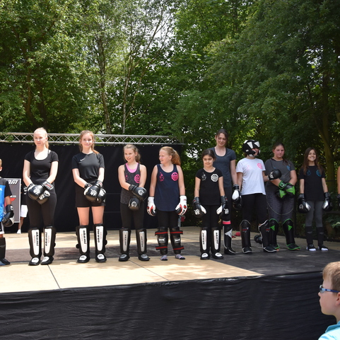Gruppe von Kindern und Jugendlichen in Sportkleidung mit Boxhandschuhen auf einer B&uuml;hne im Freien.