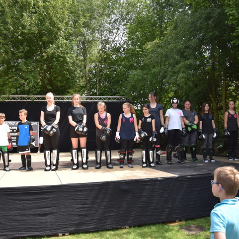 Gruppe von Kindern und Jugendlichem in Boxausr&uuml;stung steht auf einer B&uuml;hne im Freien, umgeben von Zuschauern.