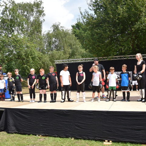 Kinder in Sportbekleidung stehen auf einer B&uuml;hne, w&auml;hrend Erwachsene im Hintergrund zuschauen. Gr&uuml;ne B&auml;ume im Hintergrund.