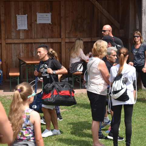 Menschengruppen stehen auf einer Wiese, w&auml;hrend sich einige miteinander unterhalten und Taschen halten.