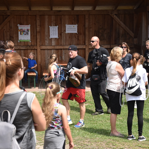 Menschenmenge vor einer Holzstruktur, ein Mann in rot-schwarzer Kleidung tr&auml;gt Ausr&uuml;stung, das Gras ist gr&uuml;n.