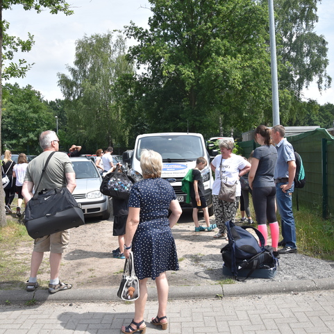 Gruppe von Personen mit Gep&auml;ck wartet an einer Landstra&szlig;e neben parkenden Autos und einem Lieferfahrzeug.