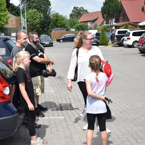 Gruppe von f&uuml;nf Personen steht auf einem Parkplatz neben mehreren Autos, es ist sonnig und gr&uuml;n um sie herum.